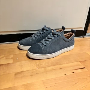 Blå mockasneakers från Christian Louboutin - Snygga blå sneakers i mocka från Christian Louboutin med klassisk vit sula och den ikoniska röda undersidan. Skorna har rund tå, snörning och diskreta detaljer. Insidan är beige och de har en stilren, minimalistisk look som passar till många outfits. Säljes pga att dem är för små kan inte garantera äkthet då dem är köpta här på plick därför de säljes billigt. Box och dustbag medföljer