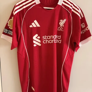 Liverpool Salah 11 matchtröja Adidas röd - Liverpool FC matchtröja i rött med vita detaljer, Adidas-logga och klubbmärke broderat på bröstet. Nummer 11 och storlek L. Salah på ryggen, Premier League-märke på ärmen. Tillverkad i ventilerande polyester, kortärmad och med klassiska ränder på axlarna.
