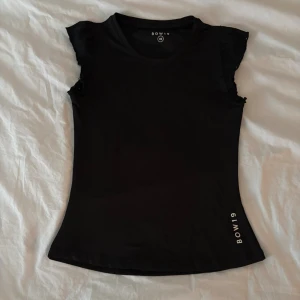 Svart topp med volangärm från BOW19 - Svart figurnära topp från BOW19 i storlek XS. Toppen har korta volangärmar och rund halsringning. Materialet känns mjukt och stretchigt, perfekt för en stilren look och till träning. Diskret BOW19-tryck nertill och i nacken. Perfekt för en snygg träningsoutfit💕