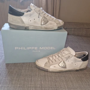 Philippe Model vita sneakers strl 44 - Säljer ett par vita Philippe Model sneakers i skinn med grå och svarta detaljer. Skorna har snörning med logotyp på skosnörena och en silverfärgad sköld på sidan. Klassisk låg modell med rund tå och platt sula. Tillverkade i Italien.
