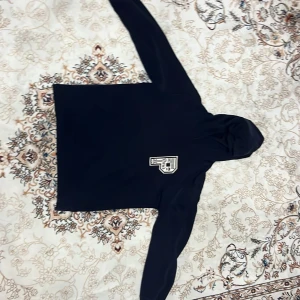 Svart hoodie med Paris-tryck - Svart hoodie med stor huva och tryck på bröstet samt texten 'PARIS' i vitt på ryggen. Klassisk modell med långa ärmar och mjukt material, perfekt för en chill och stilren look.