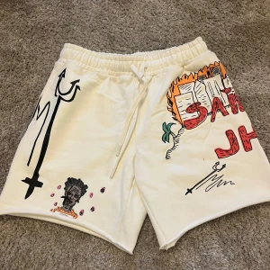 Vita bomullsshorts med grafiska tryck - Unika vita shorts i 100% bomull med elastisk midja och snörning. Shortsen kommer från Christian Sex Club. Ord. Pris 1040 kr. Shortsens framsida och baksida är täckta av coola grafiska tryck, teckningar och text, bland annat eld, palmer, ansikten och Brooklyn- och musikmotiv. Perfekta för dig som vill sticka ut med streetstyle. 