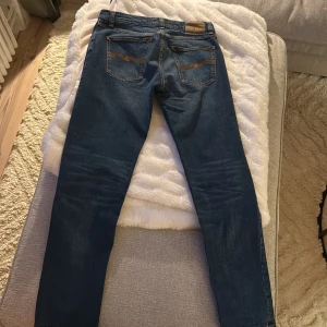 Blå jeans från Nudie Jeans W30 L32 - Säljer ett par klassiska blå jeans från Nudie Jeans i modellen W30 L32. Jeansen har fem fickor, orangea sömmar och en snygg tvätt. Tillverkade i slitstarkt bomullsmaterial och har en rak passform med knappgylf. Perfekta för dig som gillar stilrena och tidlösa jeans.