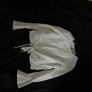 Vit omlottopp med knyt och struktur - Säljer en vit omlottopp från Monki med lång ärm och knyt i midjan. Toppen har en strukturerad, lätt skrynklig yta och vida ärmslut som ger en cool look. Perfekt för dig som gillar stilrena plagg med en twist.