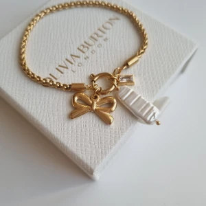 Guldarmband med rosett  - Charmigt guldarmband med en söt rosettberlock, en vit pärlberlock och en liten glittrig detalj. Kedjan är tvinnad och har en elegant finish som ger en lyxig känsla. Perfekt för dig som gillar detaljer och vill sticka ut. Lådan ingår ej.