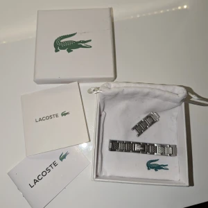 Silverfärgat armband från Lacoste - Stilrent armband från Lacoste i 925 silver med klassisk krokodil-logga. Länkarna har ett snyggt mönster och armbandet kommer i originalask med tillhörande påse och papper. Perfekt accessoar för dig som gillar sportig och modern stil. Går förminska och förstora med extra metall biten som följer med. MVH