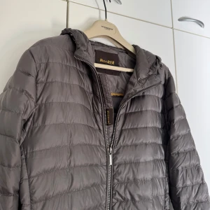 Grå dunjacka från MooRER med huva - Snygg grå dunjacka från MooRER med huva och quiltad design. Jackan har dragkedja framtill och är fylld med 100% gåsdun för maximal värme. Perfekt för dig som vill ha en stilren look och samtidigt hålla dig varm under kyliga dagar. Har en liten defekt vid ärmen, men den syns inte vid användning
