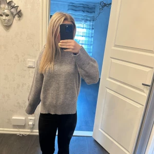 Grå stickad tröja med rund hals - Mysig grå stickad tröja med rund halsringning och breda ribbade muddar vid ärmslut och nederkant. Tröjan har en loose passform och är perfekt för kyligare dagar. Enkel och stilren design som är lätt att matcha med olika outfits.