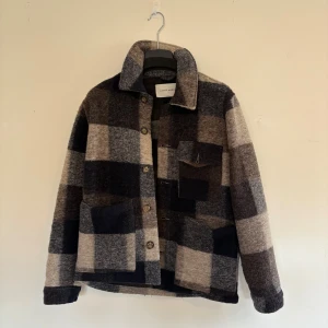 Rutig overshirt från A Day's March - Snygg overshirt från A Day's March i rutigt mönster med stora rutor i mörkblått , grått och beige. Jackan har klassisk krage, knäppning framtill och tre fickor. Tillverkad i mjuk ullblandning, perfekt för lager-på-lager under höst och vinter.
