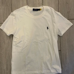 Vit t-shirt från Polo Ralph Lauren - Klassisk vit t-shirt från Polo Ralph Lauren med broderad svart logga på bröstet. T-shirten har rund halsringning och korta ärmar. Tillverkad i mjuk bomull för en skön och stilren look. Jag har sytt ihop lapparna på insidan eftersom de gick av efter jag slet upp tröjan när den var ny😬 men annars är den i nytt skick eftersom jag inte använt den nånting sen jag fick den! Skriv för fler bilder!!