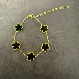 Guldarmband med svarta blommor - Snyggt armband med sex svarta blommor i fyrklöverform, infattade i guldfärgad kedja. Blommorna har en fin, pärlad kant och armbandet har justerbar längd med karbinlås. Perfekt accessoar för att lyfta din stil.