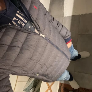 Grå pufferjacka från Polo Ralph Lauren - Säljer en grå pufferjacka från Polo Ralph Lauren med röd logga på bröstet. Jackan har quiltad design, dragkedja framtill och två sidofickor med dragkedja. Ända defekten är ett litet hål men det syns knappt och är inte till besvär . Perfekt för kyliga dagar! 