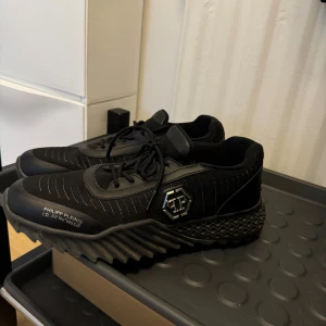 Philipp Plein svarta sneakers - Svarta sneakers från Philipp Plein med coola detaljer som silverfärgad logotyp på sidan och dödskalle på hälen. Skorna har snörning, mönstrad sula och är tillverkade i en mix av textil och skinn. Snygga linjer och modern design gör dem till ett statement.