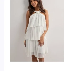 Vit volangklänning med halterneck - Superfin vit kortklänning med halterneck och flera lager volanger. Klänningen har en flowy känsla och är ärmlös, perfekt för sommarens alla tillfällen. Snygg knutdetalj vid halsen och lätt, luftig passform.