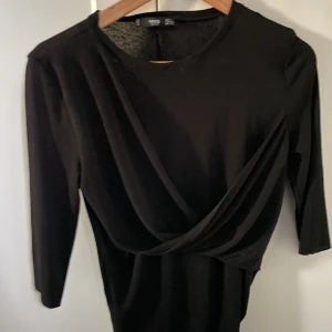 Svart omlottopp från Mango - Fin svart tröja från mango med fin applikation vid bröstet! Sitter tajt och är tre kvart ärmad!
