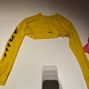 Gul croppad långärmad Nike-tröja - Gul croppad långärmad tröja från Nike med svart logga på ryggen och stort Nike-tryck längs ena ärmen. Tröjan har en loose fit som ger en avslappnad vibe. Perfekt för dig som gillar sportig och trendig stil.