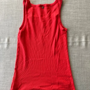 Röd ribbad tanktop i bomull från Boss Orange  - Cool röd tanktop från Boss Orange med ribbad struktur och rundad hals. Toppen är ärmlös och har en liten metall-logga bak samt ett märke fram. Perfekt för dig som gillar enkel och stilren design i stark färg.