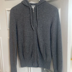 Grå hoodie i kashmir från Soft Goat - Mysig grå hoodie från Soft Goat i kashmir. Tröjan har dragkedja framtill, huva med snörning och två fickor. Perfekt för dig som gillar en stilren och enkel look med premiumkänsla.