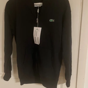 Svart kofta från Lacoste  - Svart hoodie från Lacoste med klassisk krokodillogga på bröstet. Tröjan har half zip, huva och en stor magficka. Perfekt för dig som gillar stilrena och sportiga plagg. Materialet är mjukt och skönt, troligtvis bomull eller bomullsblandning.