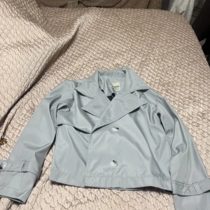 Ljusgrå kort trenchcoat - Snygg ljusgrå kort trenchcoat från Vero Moda med dubbelknäppning och klassisk krage. Jackan har långa ärmar med knäppdetalj vid ärmslut och är tillverkad i ett slätt, syntetmaterial. Perfekt för dig som vill ha en stilren och trendig look.