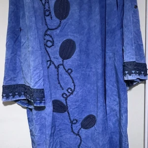 Blå långklänning med broderi - Unik blå långklänning med dekorativa broderier och pärldetaljer framtill. Klänningen har vida ärmar med snygga kantdetaljer och ett mjukt, lätt material. Perfekt för dig som gillar bohemisk och konstnärlig stil.