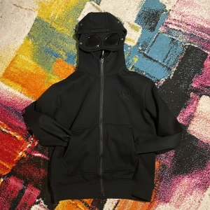 Svart gogglejacka från C.P. Company - Svart jacka från C.P. Company med ikonisk goggle-hood och hel dragkedja framtill. Jackan har två stora fickor, broderad logga på bröstet och är tillverkad i ett mjukt, stretchigt material. Perfekt för dig som vill sticka ut med en unik streetwear-look.   Hoodien är en m men sitter som en small. Tar även byten