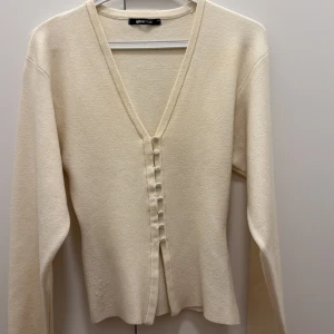 Vit kofta från Gina Tricot - Snygg vit kofta från Gina Tricot med djup v-ringning och klädda knappar framtill. Långärmad och i mjukt material som känns skönt mot huden. Perfekt för lager-på-lager och enkel att matcha med olika outfits.