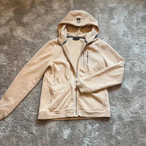 Beige hoodie från Sail Racing - Snygg beige hoodie från Sail Racing med dragkedja framtill och på bröstfickan. Tröjan har huva med broderad logga och är tillverkad i mjukt material. Perfekt för dig som gillar stilren och sportig look.Helt oanvänd.