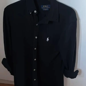 Mörkblå skjorta från Polo Ralph Lauren XS - Svart långärmad skjorta från Polo Ralph Lauren i lätt mesh-bomull. Klassisk krage, vita knappar framtill och ikonisk broderad logga på bröstet. Perfekt för dig som gillar stilrena och tidlösa plagg med snygg passform.