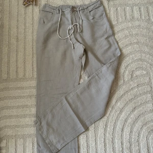 Beiga linnebyxor med snörning - Snygga beiga linnebyxor med raka ben och snörning i midjan. Perfekta för en chill och avslappnad stil. Byxorna har klassiska fickor fram och bälteshällor för extra detalj.