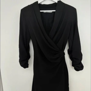 Svart omlottklänning med knyt - Stilren svart kortklänning med omlott-design och knyt i sidan. Klänningen har långa ärmar och djup v-ringning framtill. Materialet är mjukt och följsamt, perfekt för en trendig look. Snygg detalj med drapering vid midjan och bälte baktill.
