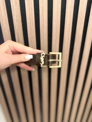 Ingen väst på bilden - Bilden visar ett brunt bälte med guldfärgat spänne och YSL-detalj, ingen väst syns.