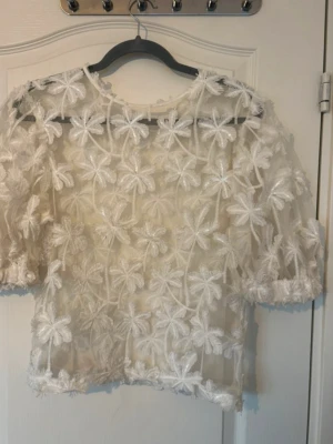 Vit blommig meshblus från Object - Säljer en superfin vit meshblus från Object i storlek 34. Blusen är transparent med stora broderade blommor över hela och har halvlånga puffärmar. Dragkedja finns på ärmen för extra detalj. Perfekt för dig som vill sticka ut lite extra!