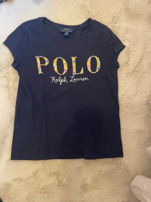Mörkblå t-shirt från Polo Ralph Lauren - Snygg mörkblå t-shirt från Polo Ralph Lauren med färgglatt blommigt tryck på bröstet och vit broderad text. Klassisk rund hals och korta ärmar. Tillverkad i mjuk bomull som känns skön mot huden. Perfekt till jeans eller shorts.                   Storlek: Medium kids, 136-142 cm