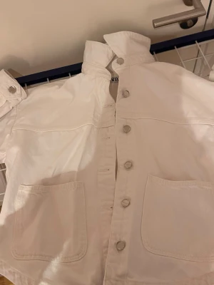 Vit jeansjacka från Zara XS - Säljer en vit jeansjacka från Zara i storlek XS. Jackan har stora framfickor, knappar framtill och vid ärmsluten samt en hög krage. Perfekt för dig som gillar en clean och stilren look.