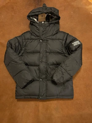 Svart pufferjacka från Burberry - Säljer en svart pufferjacka från Burberry med huva och klassiskt rutigt foder. Jackan har både dragkedja och knappar framtill samt en patch med Burberry London England på ärmen. Perfekt för kalla dagar och riktigt snygg till streetwear.