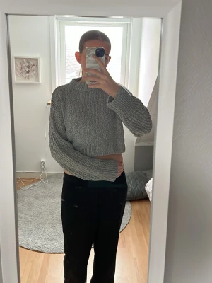 Grå croppad stickad tröja - Säljer en grå croppad stickad tröja med lång ärm och högre hals. Tröjan har en loose fit och är perfekt för lager-på-lager-stil. Enkel och stilren design som passar till många outfits.
