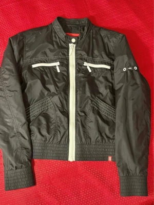 Svart bomberjacka från edc by Esprit - Snygg svart bomberjacka från edc by Esprit med vita dragkedjor och detaljer. Jackan har två bröstfickor med dragkedja, ribbade muddar och krage samt coola sömmar. Tillverkad i glansig polyester som ger en sportig vibe.