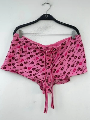 VS Rosa shorts med gitarrmönster - Säljer ett par rosa shorts med coolt gitarrmönster i mörkrosa och brunt. De har snörning i midjan och är tillverkade i mjukt bomullstyg. Perfekta för chill dagar eller som pyjamasshorts. Passformen är loose och de har en avslappnad vibe. Storleken är L men passar mig som brukar ha S/M
