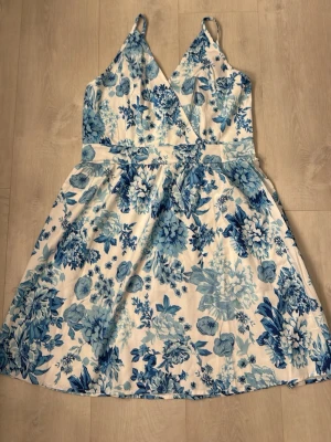 Vit och blå blommig kortklänning - Supersöt kortklänning med vita och blå toner och ett blommigt mönster. Klänningen har smala axelband, omlott upptill och markerad midja. Tillverkad i ett lätt och svalt material, perfekt för varma dagar. Dragkedja i sidan för enkel påklädning.