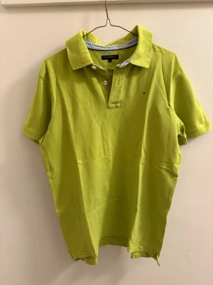 Limegrön piké från Tommy Hilfiger - Fräsch limegrön pikétröja från Tommy Hilfiger med klassisk krage och två knappar framtill. Skjortan har korta ärmar och diskret logga på bröstet. Tillverkad i mjuk bomull som känns skön mot huden. Perfekt för dig som vill sticka ut med färg.