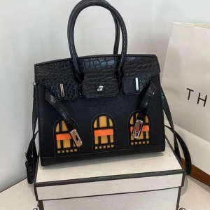 Svart handväska med grafiskt mönster - Cool svart handväska med krokodilpräglad överdel och grafiskt fönstermönster i gult och orange på framsidan. Väskan har metallspännen, dubbla handtag och axelrem. Perfekt för dig som vill sticka ut med en unik design.