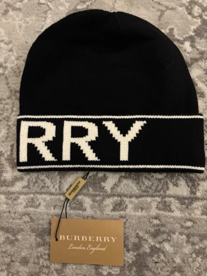 Svart mössa från Burberry - Säljer en svart mössa från Burberry med bred vit kant och stor logotyp i vitt runt om. Klassisk och stilren design med ribbstickad känsla och mjukt material. Perfekt accessoar för dig som vill ha något trendigt och exklusivt.