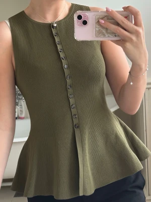 Olivgrön ribbad ärmlös topp med knappar - Säljer en olivgrön ribbad topp med utsvängd nederdel och dekorativa knappar framtill. Toppen är ärmlös och har en rund halsringning. Perfekt att styla med både jeans och kjol för en snygg look.