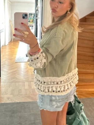 Grön jacka Zara  - En trendig grön jacka från Zara som inte säljs längre med unika virkade detaljer och tofsar i vitt längs nederkant och ärmslut. Jackan har knappar är i super skick