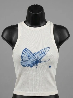 Vit croppad linnetopp med blå fjäril - Säljer en vit croppad linnetopp med ribbad struktur och ett stort blått fjärilstryck framtill. Toppen har breda axelband och rund halsringning, perfekt för en trendig och avslappnad look.