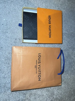 Stilren svart plånbok i monogrammönster - Stilren svart plånbok i monogrammönstrad coated canvas. Klassisk och tidlös modell med flera kortfack och sedelfack – både praktisk och elegant.  I fint skick med normala bruksspår (se bilder). Originalbox medföljer. Påse ingår ej.