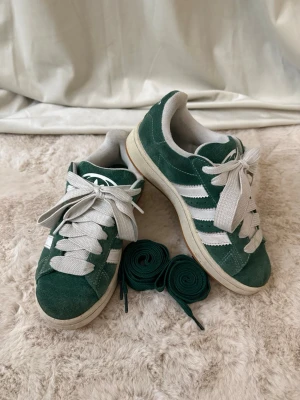 Adidas Campus gröna sneakers mocka - Gröna Adidas Campus sneakers i mocka med vita detaljer och klassiska tre ränder på sidan. Skorna har vit sula, breda vita skosnören och extra gröna skosnören medföljer. Adidas-logga på plös och häl. Låg modell med rund tå och platt sula.