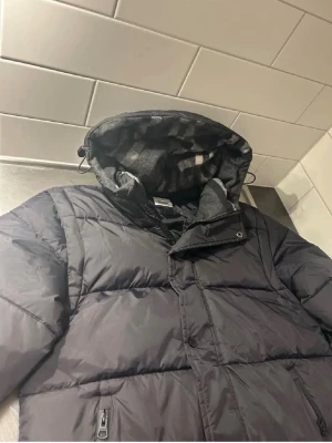 Svart pufferjacka från Burberry - Säljer en svart pufferjacka från Burberry London England i storlek S. Jackan har huva med mönstrat innerfoder, dragkedja och knappar framtill samt två fickor med dragkedja. Perfekt för kalla dagar och riktigt snygg streetstil.