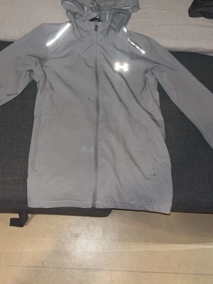 Grå Under Armour vindjacka med huva - Säljer en grå vindjacka från Under Armour med huva och hel dragkedja. Jackan har reflekterande detaljer, två sidofickor och stor logga på ryggen. Materialet är lätt och vattenavvisande, perfekt för blåsiga dagar. Storm-teknologi för extra skydd.